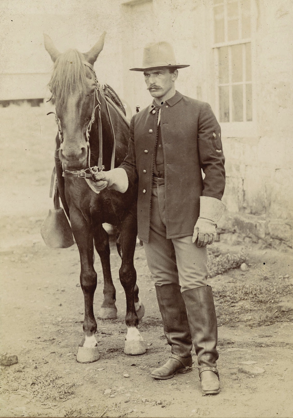 Handsome farrier wearing heavy leather gauntlets, knee-high boots, and embroidered horseshoe on shoulder, circa 1870s